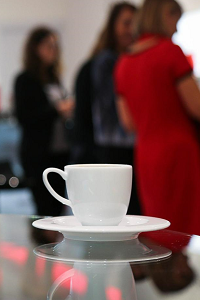 A white cup of coffee in saucer sits on a glass table while three women in the background have a discussion.
