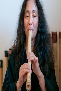 Middle aged lady with long black hair playing recorder like instrument