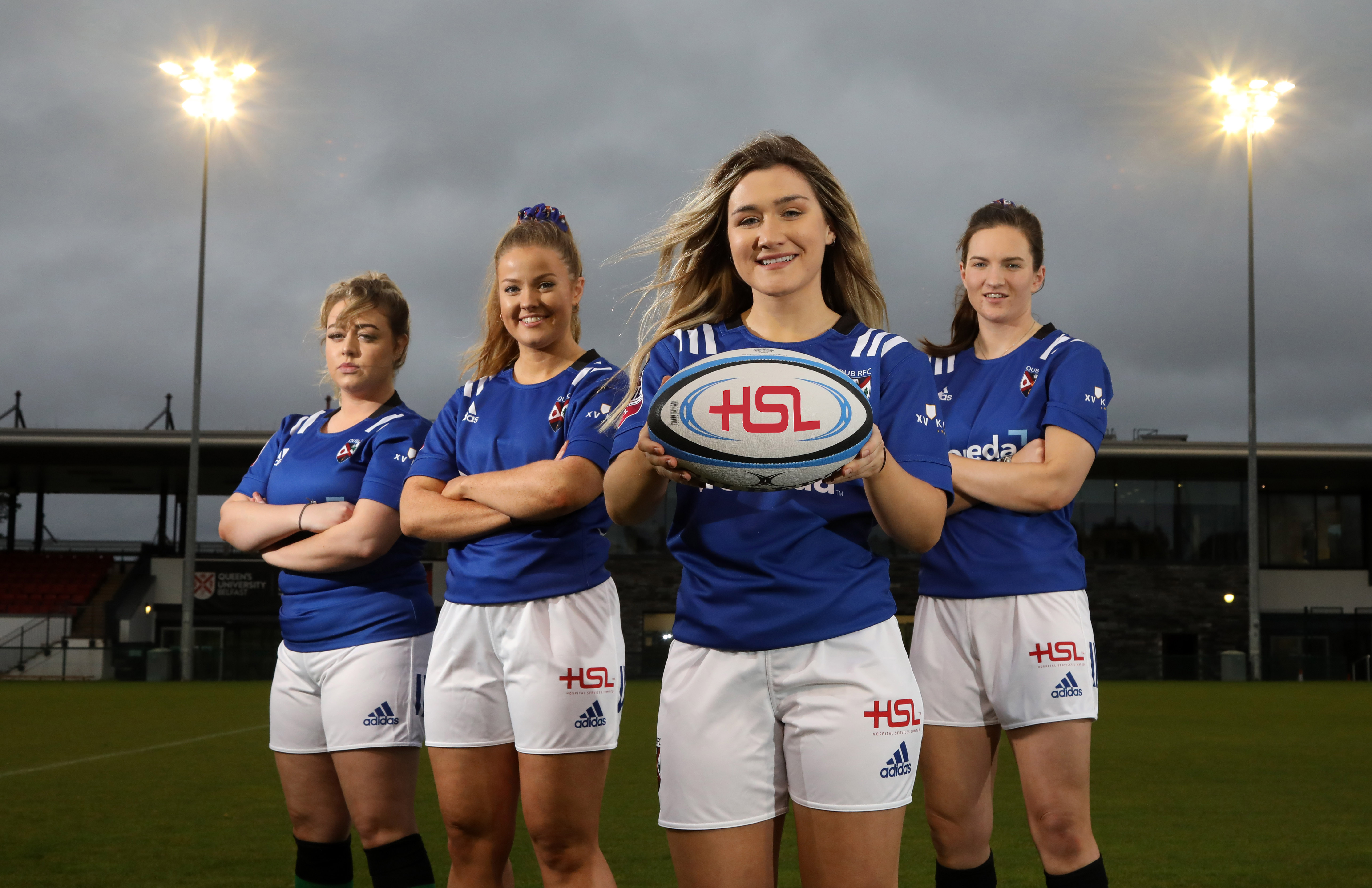 Four female rugby players stand in a row, one holding rugby ball with sponsor's initials - HSL 