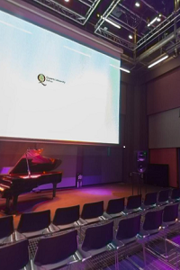 A photo of the lecture theatre of the SARC centre showing a projected image of Queen's University Belfast's logo and a piano on stage.