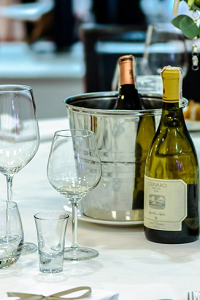 A table set with white, linen tablecloth with an ice bucket, two bottles of wine and a table set with many glasses.