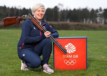 Olympic hopeful Kirsty Hegarty in Team GB tracksuit crouched in field with shotgun over her shoulder beside Team GB sign
