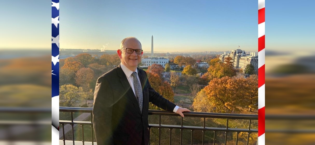 Andrew Elliott, standing with left hand on railing, with White House and Washington Monument in background, overlaid onto US flag