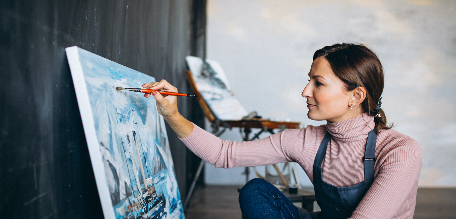 Female artist, wearing blue overalls and pink roll top jumper, with hair tied back in pony tail, painting boats and icebergs on canvas, using orange coloured paint brush 