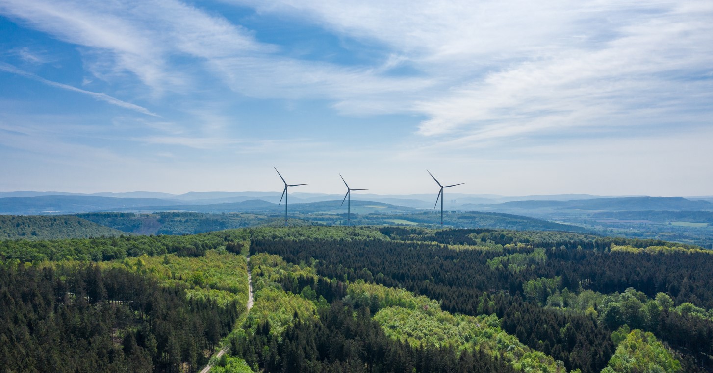 Three giant wind turbines on a hill in a forest, with blue sky behind