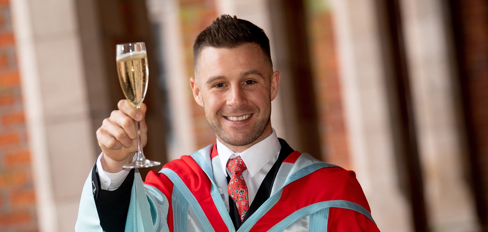Jonathan Rea in bright honorary graduate robes holding glass of champagne in cloisters