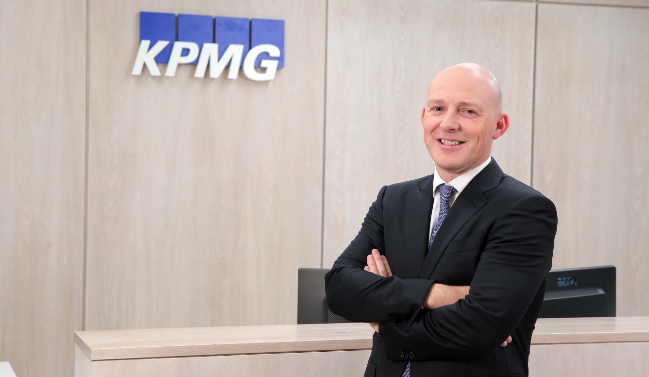 Johnny Hanna, suited with arms folded, in office in front of KPMG branded sign
