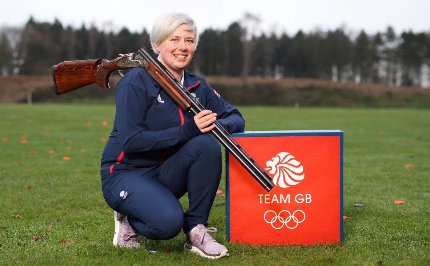 Olympic hopeful Kirsty Hegarty in Team GB tracksuit crouched in field with shotgun over her shoulder beside Team GB sign