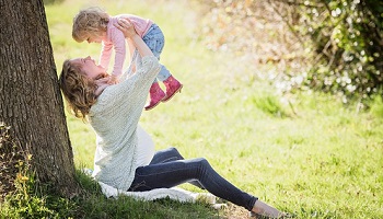 Woman holding baby aloft while seated at foot of tree in park