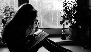 Mono image of young girl, silhouetted in front of window reading a book on her knees