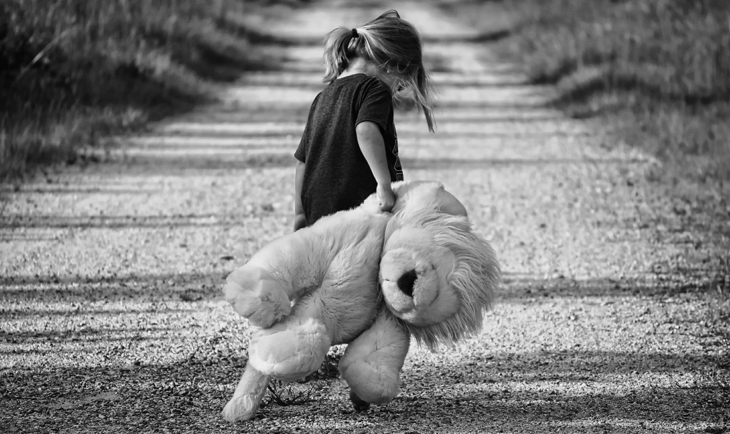 Black and white image of small girl walking on road with large teddy bear